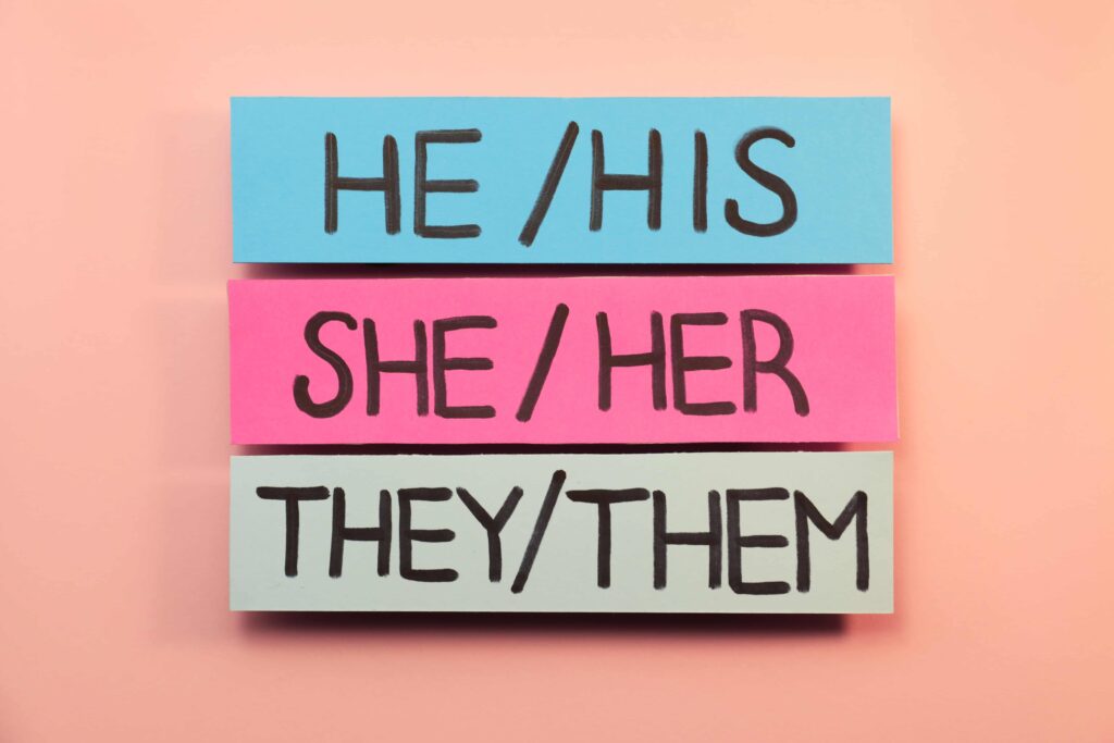 Folhas de papel com pronomes em inglês (He/His; She/Her; They/Them) em fundo rosa.