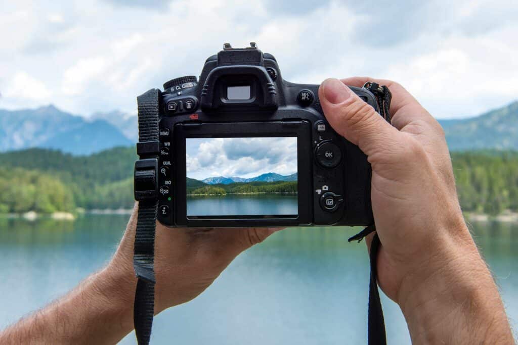 Mãos de um fotógrafo segurando uma câmera digital em frente a uma paisagem com lago e montanhas.